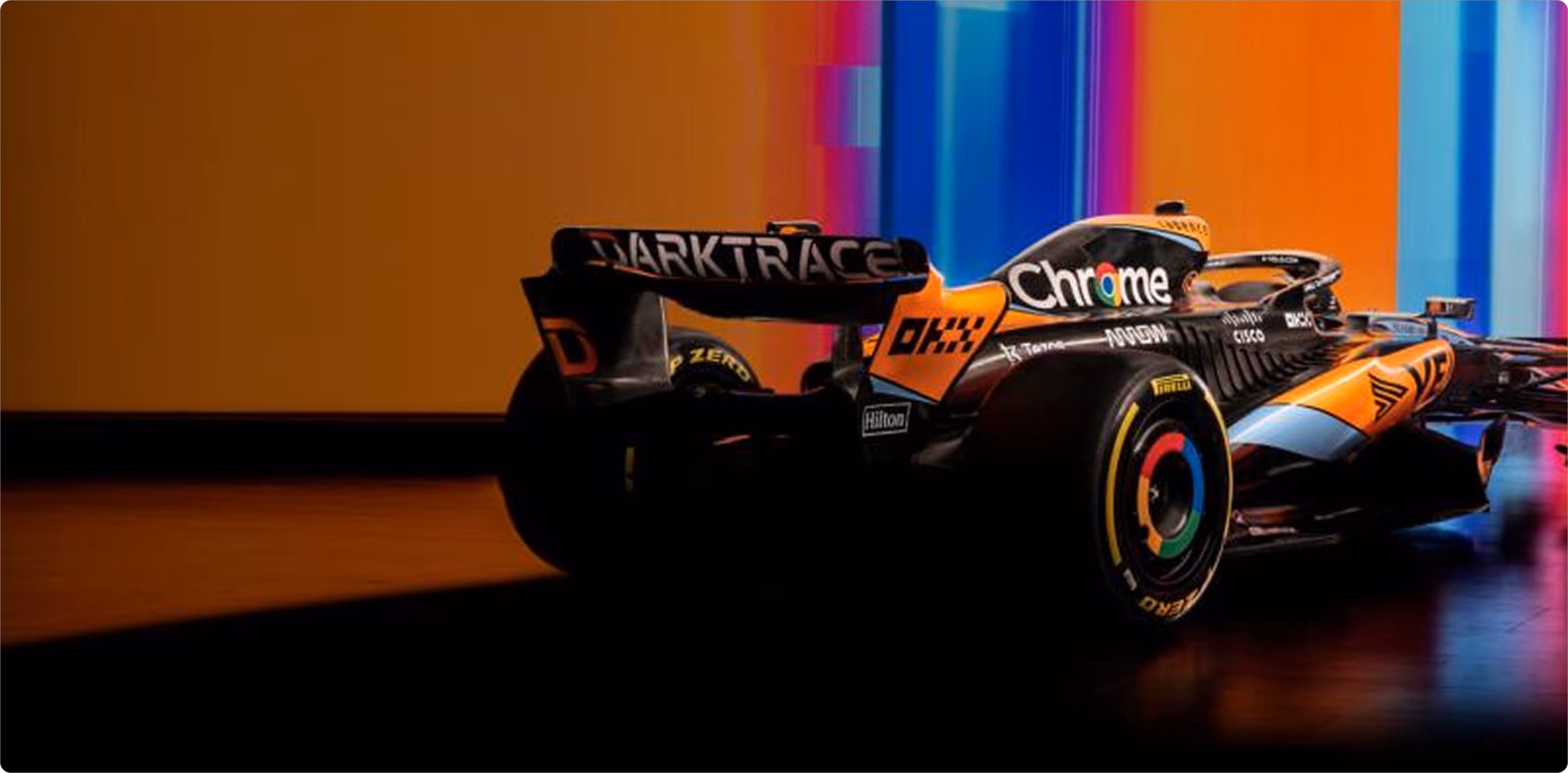 Rear side view of a black and orange Formula 1 McLaren race car with sponsor logos on a colorful background.